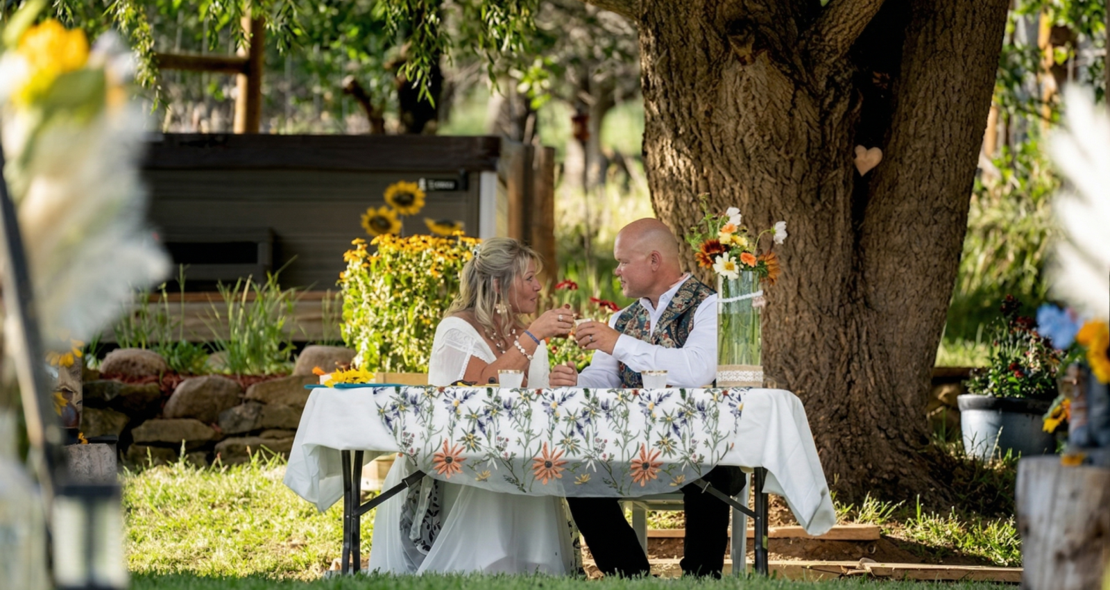 Couple enjoying an intimate outdoor wedding dinner under a tree at a rustic Colorado ranch, surrounded by flowers and natural greenery.
