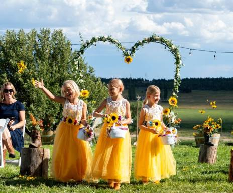 Flower girls in yellow dresses tossing petals during an outdoor wedding ceremony with a sunflower-decorated arch and scenic countryside at Three Willows Ranch in Colorado.
