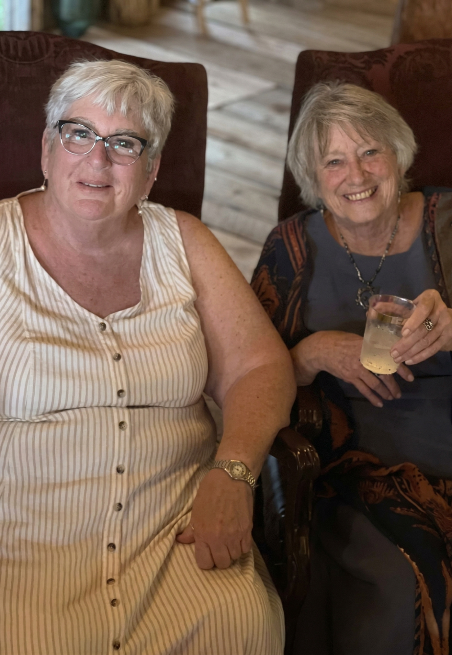 Two women smiling and relaxing with drinks while seated inside a rustic barn during a warm and welcoming ranch event.