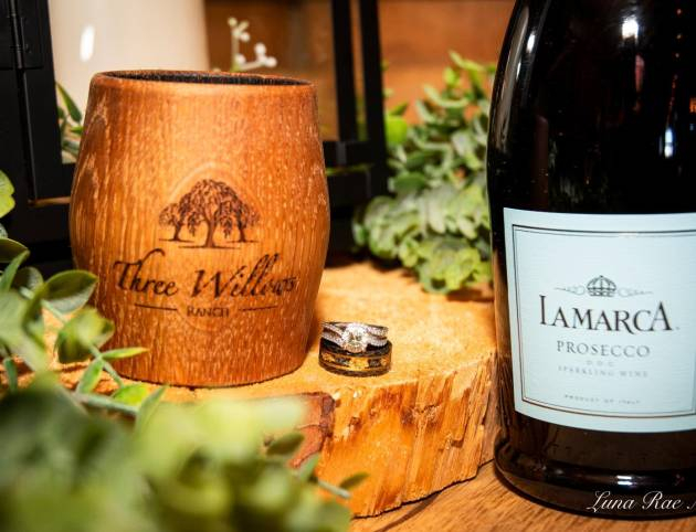 Wedding rings beside a Three Willows Ranch wooden cup and prosecco bottle on a natural wood slice, capturing elegant rustic wedding details.